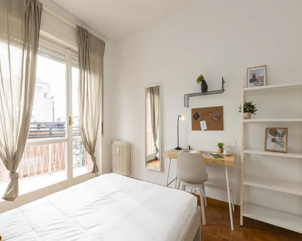 Bright, minimal bedroom with a large window, simple desk and shelving — clean linens and warm wooden floor create a welcoming space.