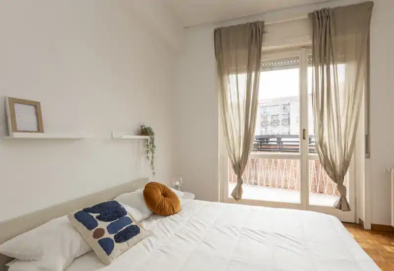 Camera da letto luminosa e arredata con un letto ben rifatto, cuscini decorativi e luce naturale morbida da una grande finestra-balcone.