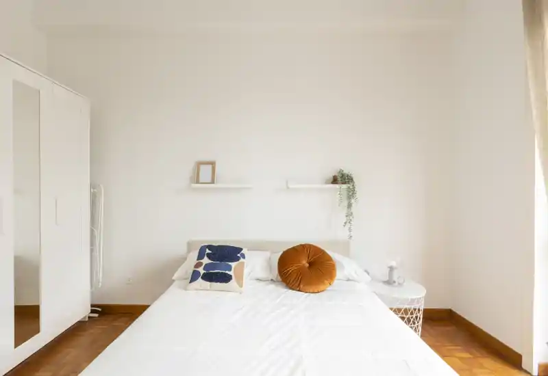Camera da letto luminosa e minimal con letto matrimoniale ben rifatto, cuscini decorativi e un piccolo comodino; la luce naturale e il pavimento in legno caldo creano un’atmosfera accogliente.