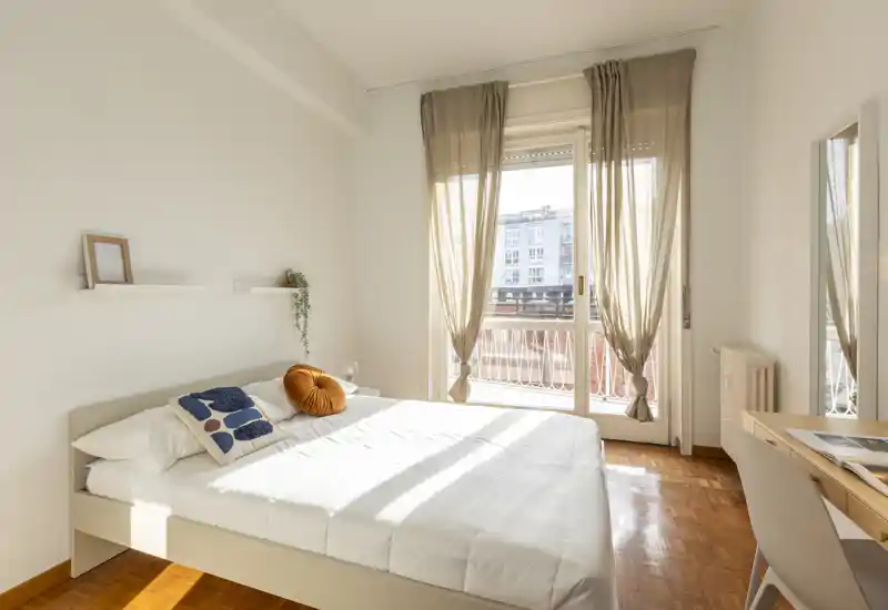 Camera da letto luminosa e minimal con letto matrimoniale, tessili morbidi e ampie porte sul balcone che lasciano entrare molta luce naturale.