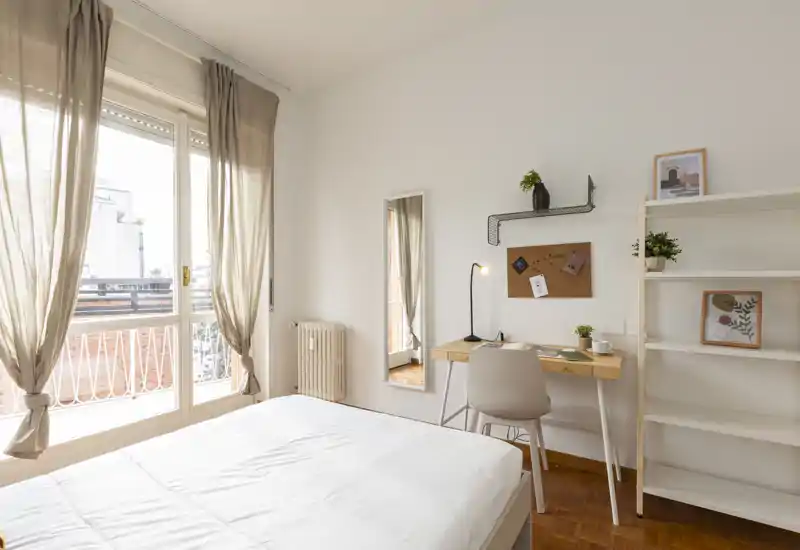 Camera da letto luminosa e minimal con ampia finestra, scrivania semplice e scaffale — biancheria pulita e pavimento in legno caldo creano un ambiente accogliente.