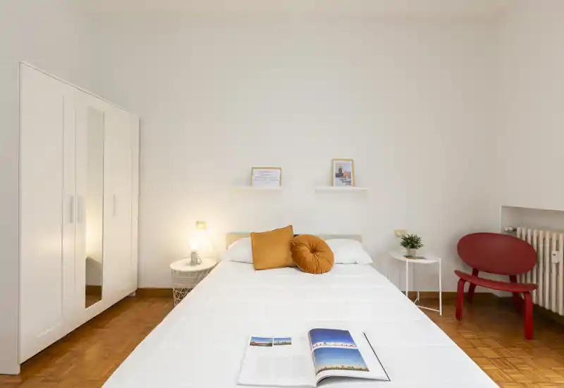 Dormitorio luminoso y minimalista con cama bien hecha, cojines cálidos, mesitas y armario — limpio, aireado y acogedor.