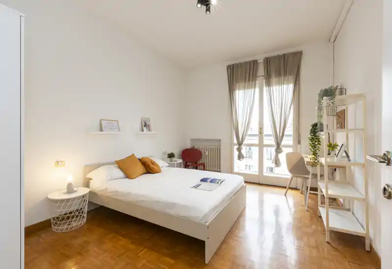 Dormitorio luminoso y ordenado con cama doble, cojines cálidos y una gran ventana que aporta luz natural. Estanterías minimalistas y un pequeño escritorio crean un espacio funcional y tranquilo.