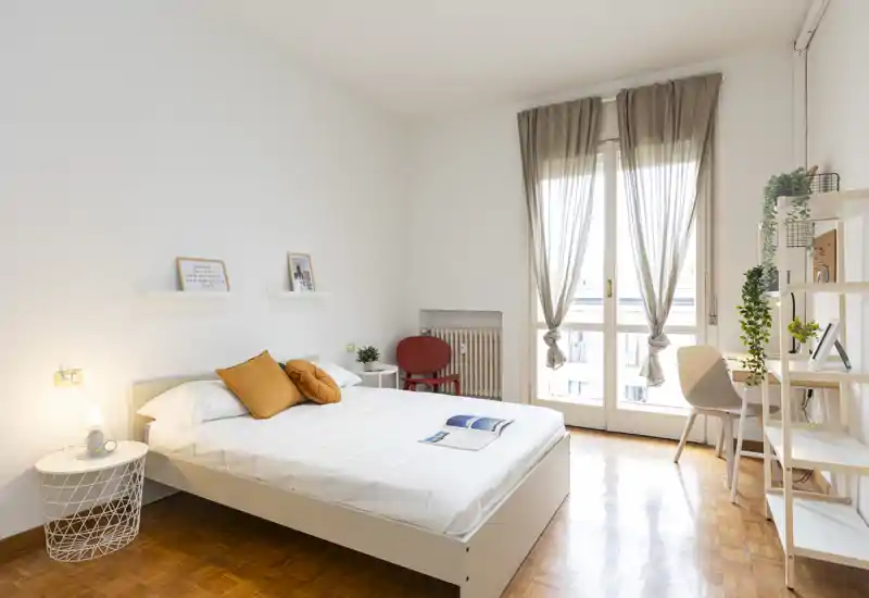 Dormitorio moderno y luminoso con cama doble, cojines cálidos y luz natural a través de las grandes puertas al balcón. Estilo minimalista, suelo de parquet y pequeño rincón de trabajo/lectura junto a la ventana.