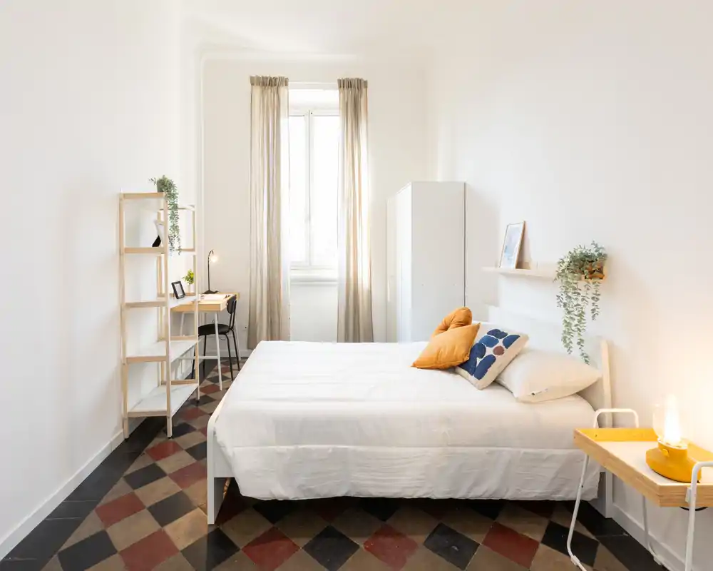 Camera da letto luminosa e minimal con letto completo in lenzuola bianche, cuscini decorativi e un piccolo comodino. Luce naturale dalla finestra centrale, scrivania compatta e scaffalatura offrono spazio funzionale e lavoro.