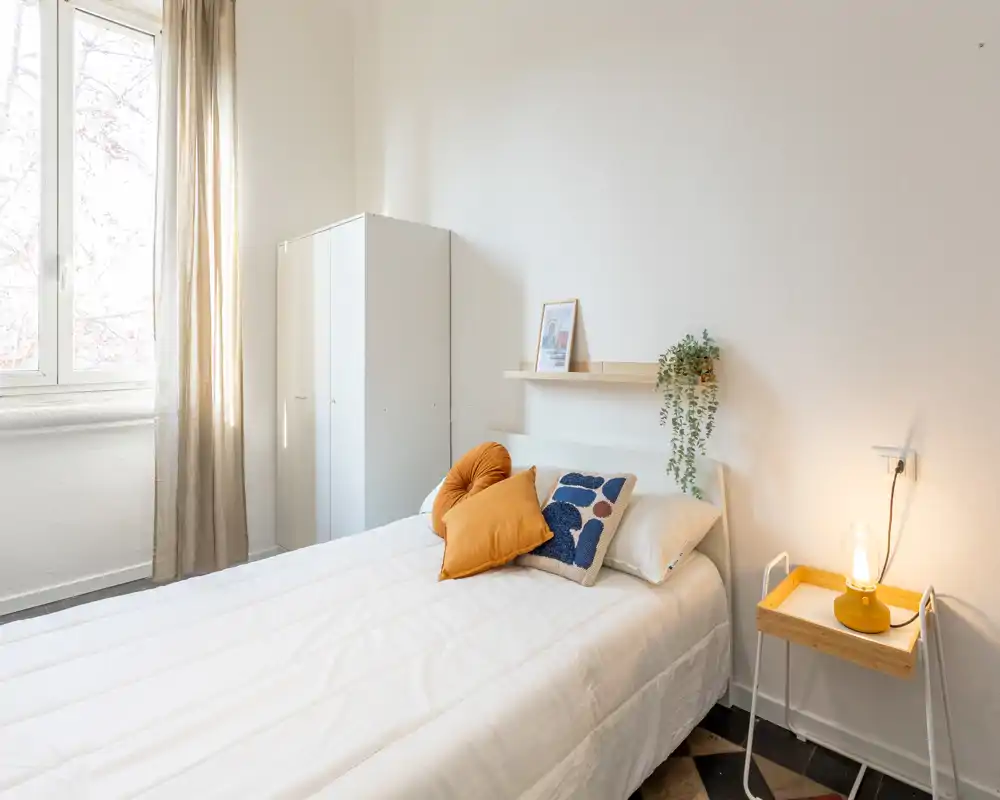 Camera singola luminosa e accogliente con letto ben rifatto, cuscini decorativi, comodino con lampada calda e luce naturale da una grande finestra.