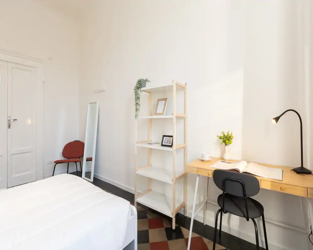 Camera da letto luminosa e minimal con letto ben rifatto, scrivania in legno con lampada e sedia e scaffalatura aperta — ideale per uno spazio di lavoro tranquillo.