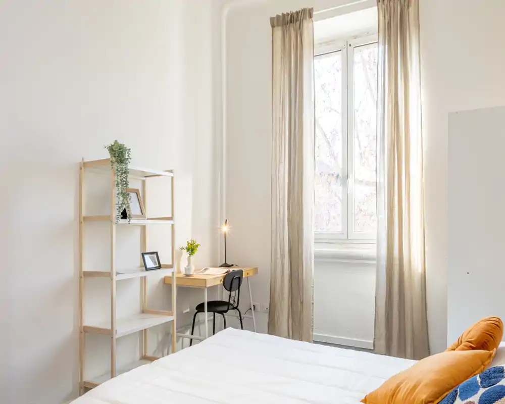 Camera da letto luminosa e ariosa con un grande letto bianco in primo piano, una scrivania minimalista vicino alla finestra e tende neutre che lasciano entrare luce naturale.