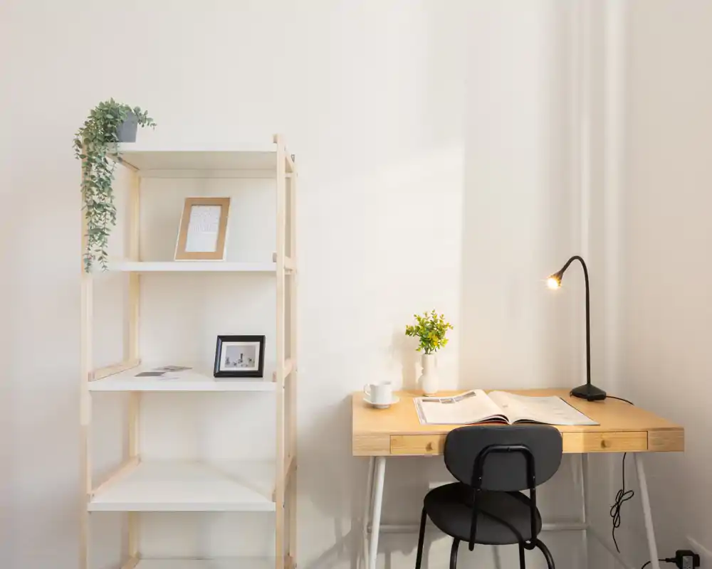 Angolo stanza in stile minimalista con scrivania in legno, scaffale e lampada da tavolo calda; ambiente luminoso, pulito e arredato in modo semplice, adatto a una camera compatta o a un angolo studio.