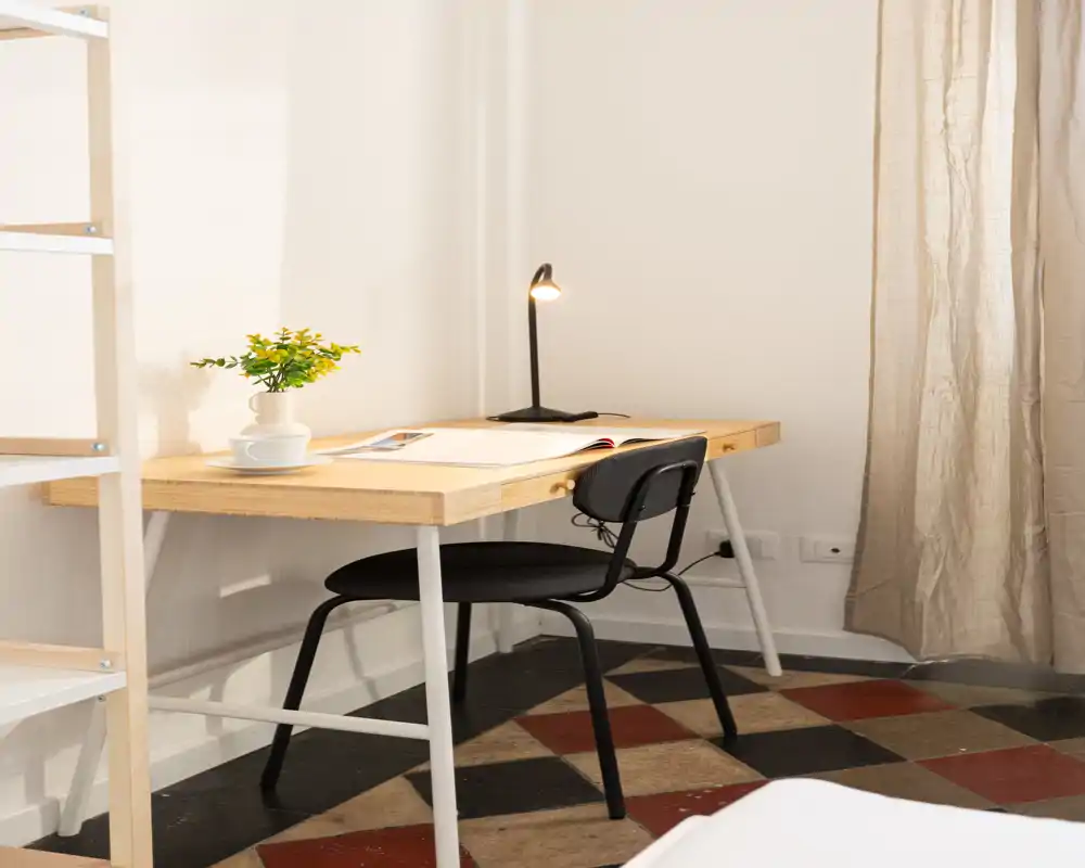 Angolo camera luminoso e minimal con una scrivania in legno e sedia nera; una piccola porzione del letto è visibile in primo piano. L'area di lavoro ha una lampada da tavolo e una piantina decorativa, perfetta per un annuncio di affitto compatto.