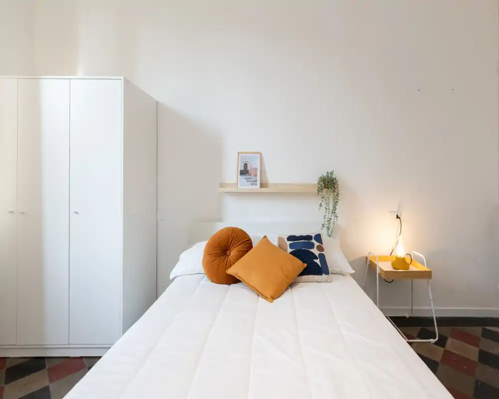 Camera da letto luminosa e moderna con letto ben rifatto, cuscini color senape, un piccolo comodino con lampada e un armadio — minimal, accogliente e ben illuminata.