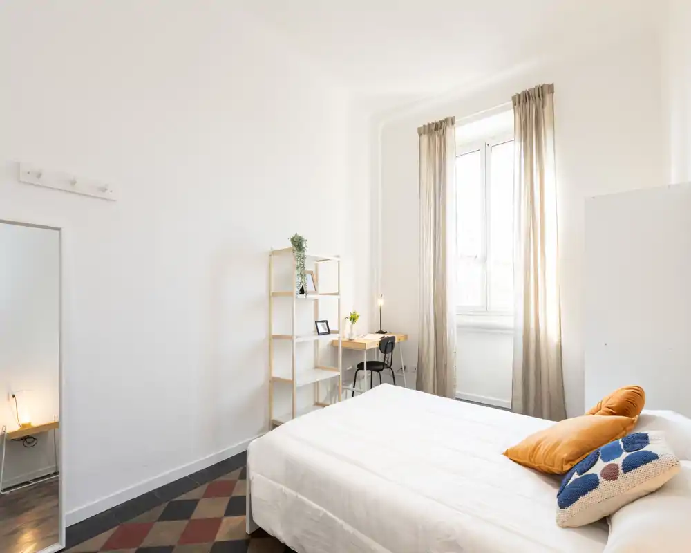 Camera da letto luminosa e minimalista con letto ben rifatto, cuscini caldi e una piccola scrivania accanto a una finestra alta che lascia entrare molta luce naturale.