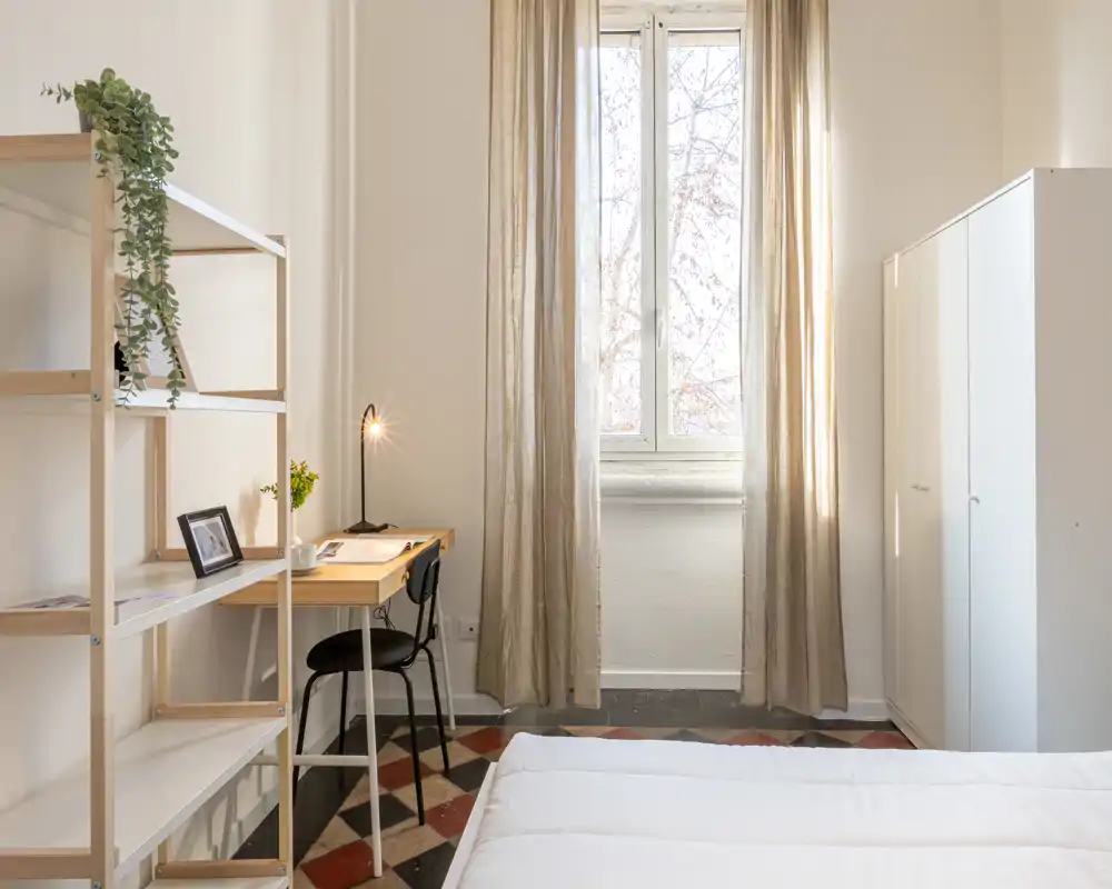 Camera da letto luminosa e minimalista con letto comodo in primo piano, scrivania e sedia vicino alla finestra e armadio bianco—ideale per un soggiorno tranquillo e adatto al lavoro.