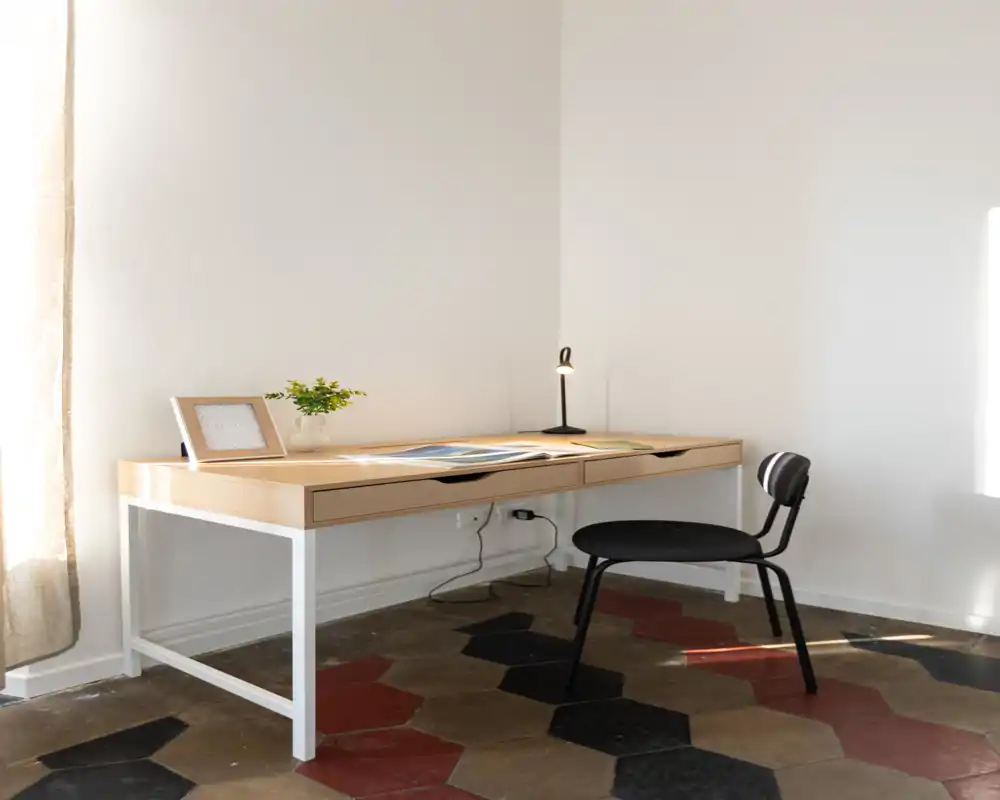 Angolo studio luminoso e minimal in una zona giorno con scrivania in legno, lampada da tavolo, sedia semplice e pianta decorativa; il pavimento piastrellato e la luce solare creano un’atmosfera ariosa e tranquilla.