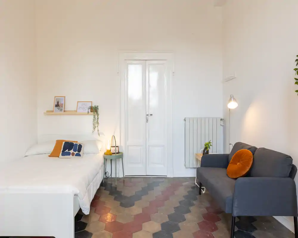 Camera da letto luminosa e stilosa con letto completo, cuscini decorativi e un angolo salotto accogliente; pavimento a piastrelle dal motivo e illuminazione soffusa creano un’atmosfera calda.
