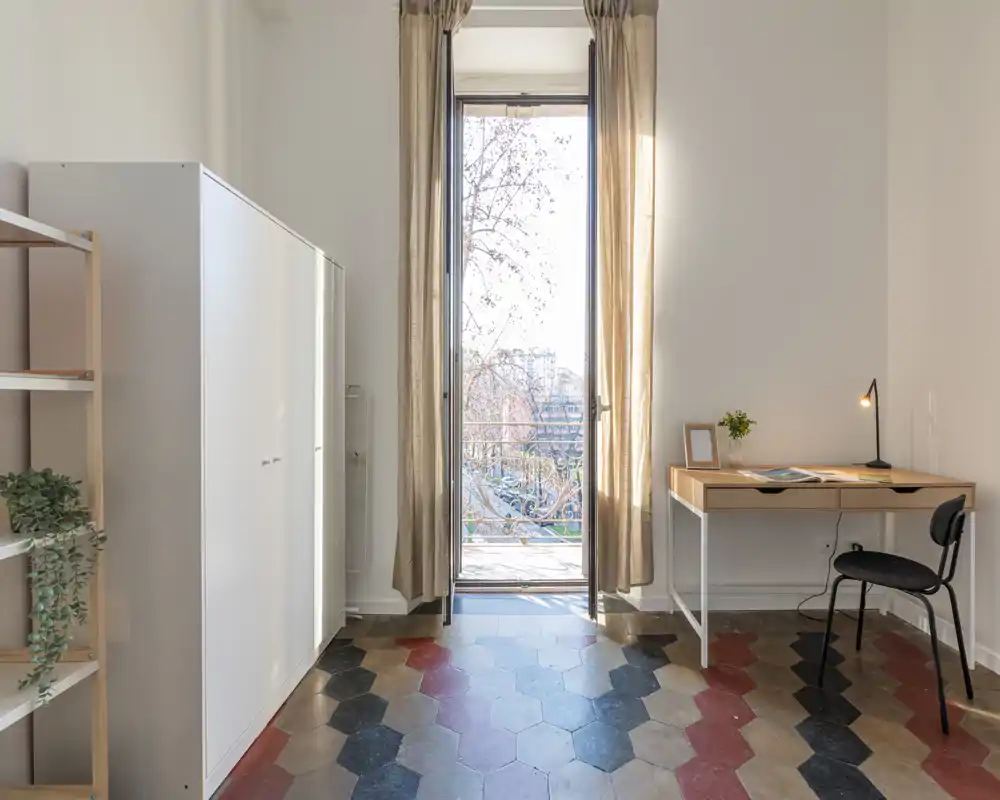 Spazio living luminoso e minimal con porta finestra sul balcone, una scrivania compatta e pavimento a piastrelle di carattere—ideale per lavoro e relax.