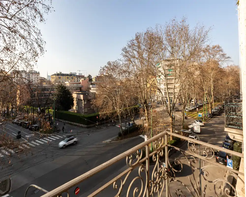 Balcone soleggiato con vista su una strada fiancheggiata da alberi e edifici residenziali, con ringhiera in ferro battuto decorativa in primo piano.