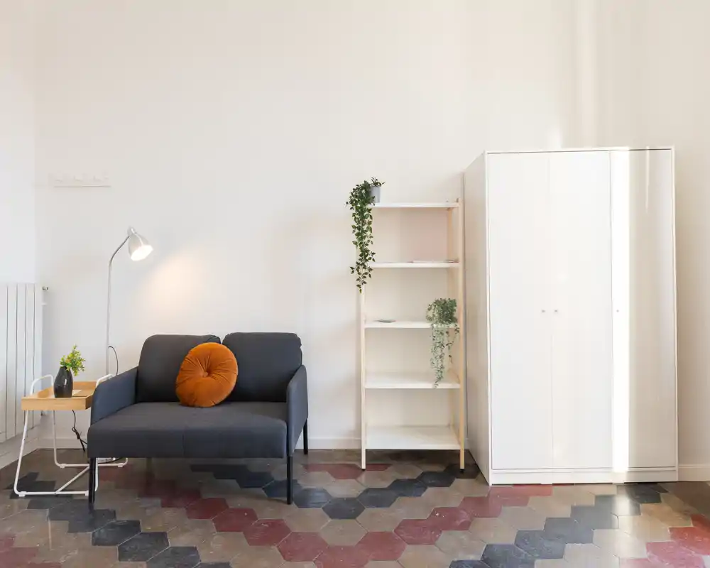 Zona giorno luminosa e minimale con divano a due posti, cuscino decorativo, lampada da terra e mensole; stile pulito in stile scandinavo con piante che rendono lo spazio accogliente.