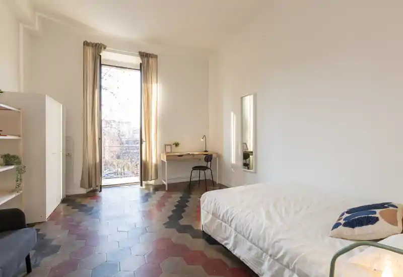 Camera da letto luminosa e ariosa con letto singolo, scrivania vicino alla finestra alta e pavimento a piastrelle esagonali decorate — ambiente pulito, minimal e soleggiato.