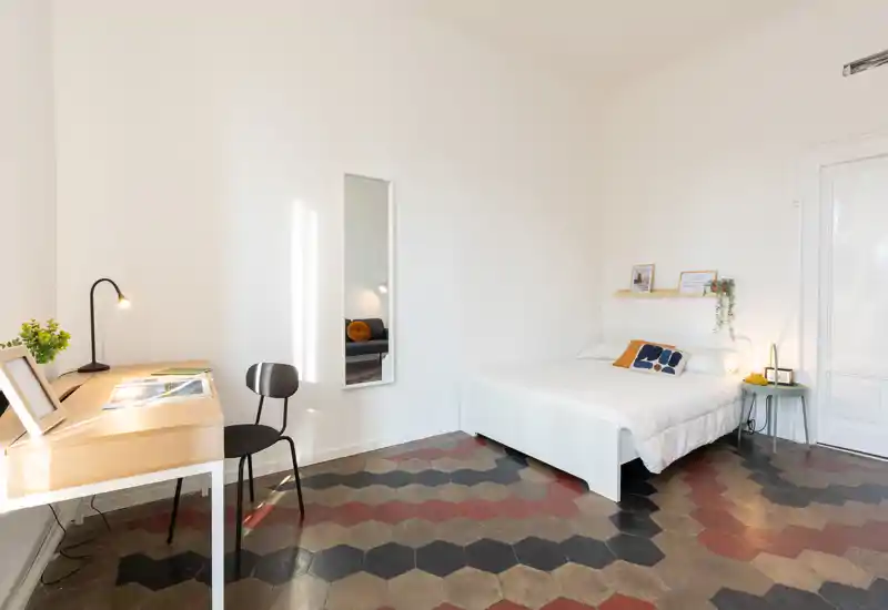 Camera da letto luminosa e minimalista con letto matrimoniale bianco, cuscini decorativi e un piccolo comodino; scrivania e sedia vicino alla finestra e pavimento a esagoni colorati che caratterizzano l'ambiente.