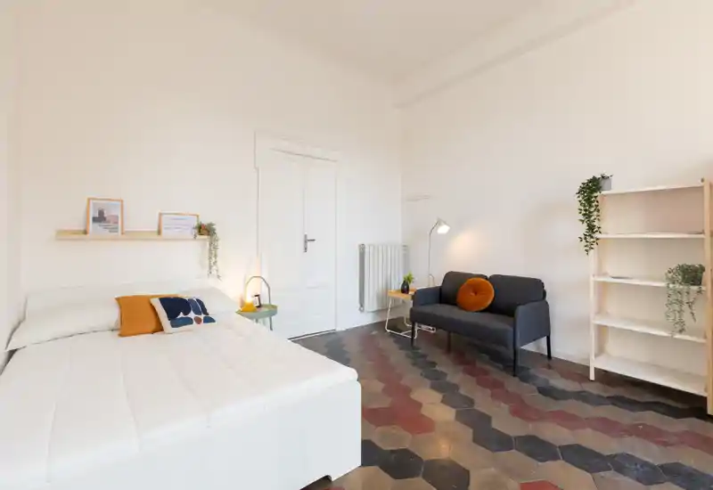 Camera da letto luminosa e di stile con letto matrimoniale, cuscini accoglienti e una piccola zona salotto con divano e lampada. Il pavimento decorato e le mensole minimal donano un aspetto moderno e arioso.