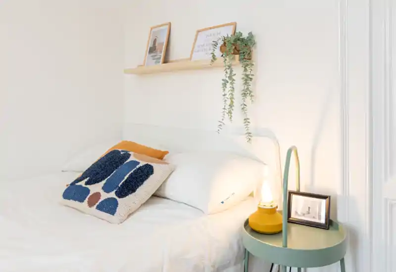 Camera da letto luminosa e minimal con letto matrimoniale ordinato, cuscini decorativi testurizzati, una mensola a muro con cornici e una pianta sospesa, e un piccolo comodino con lampada — foto perfetta per un annuncio che valorizza comfort e stile.