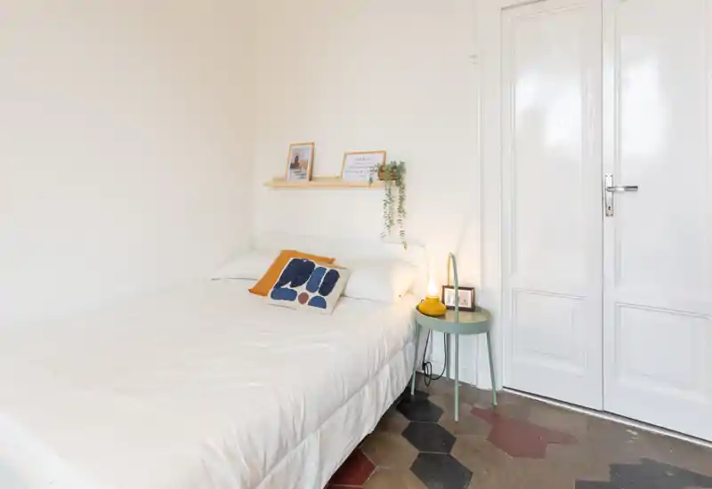 Camera da letto luminosa e minimal con letto ben rifatto, cuscini decorativi, una piccola mensola con cornici e un comodino con lampada — perfetta per annunci moderni e accoglienti.