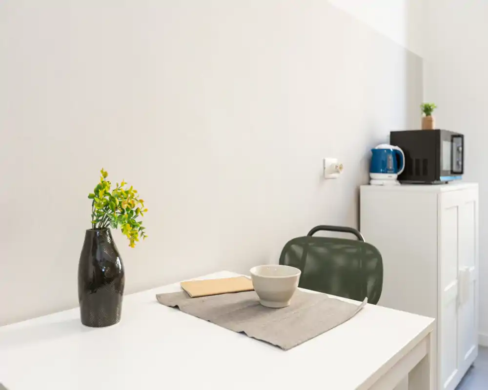 Angolo kitchenette compatto con tavolo bianco, sedia singola, piccoli elettrodomestici (bollitore e microonde) e tocchi decorativi minimi per uno spazio luminoso e funzionale.