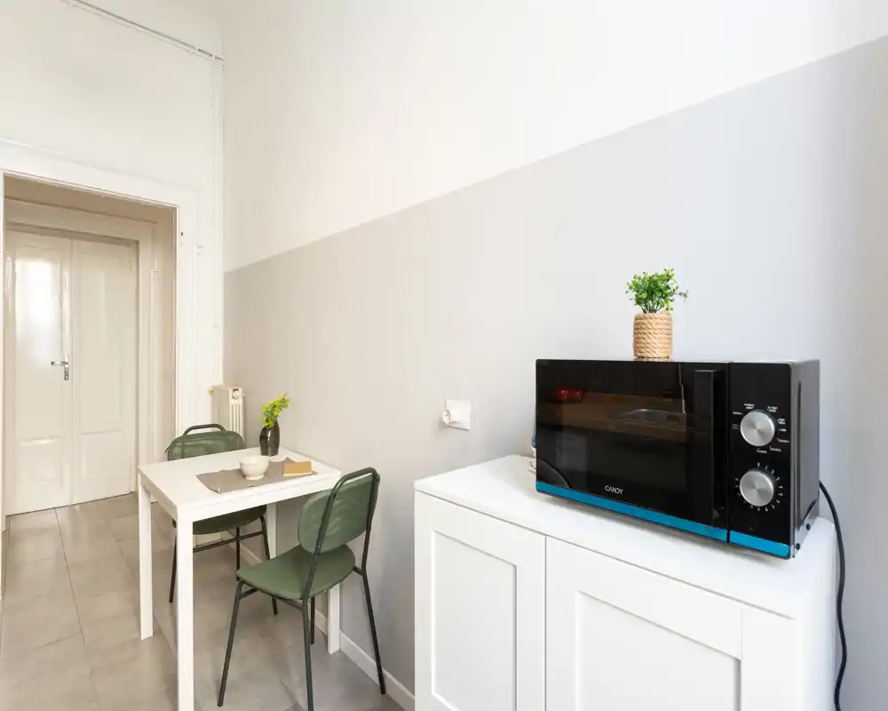 Angolo cottura compatto con piccolo tavolo da pranzo e due sedie, arredi neutri e luminosi e microonde su un mobile bianco — perfetto per monolocali o appartamenti piccoli.