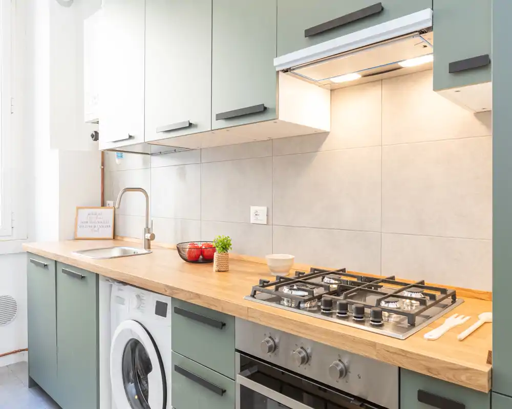 Cucina moderna e luminosa con piano di lavoro in legno, piano cottura a gas incassato, forno e lavello compatto. I mobili verde salvia, la piccola pianta e la ciotola decorativa creano un ambiente ordinato e accogliente, perfetto per annunci.