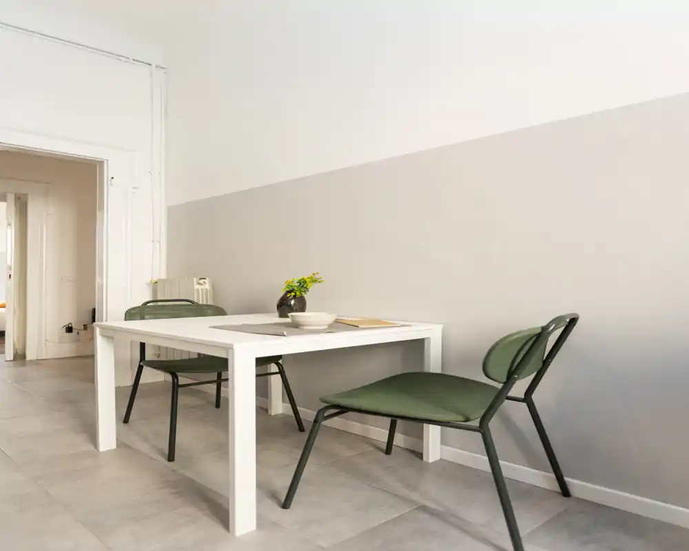 Zona pranzo minimale con tavolo bianco rettangolare e due sedie verdi, con una mise en place semplice e un vaso piccolo per un aspetto moderno e pulito.