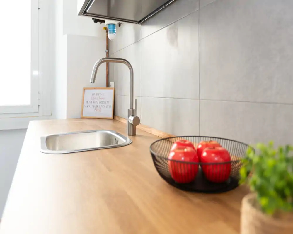 Piano cucina moderno e luminoso con lavello in acciaio e top in legno, valorizzato da una ciotola di frutta e una piccola pianta.