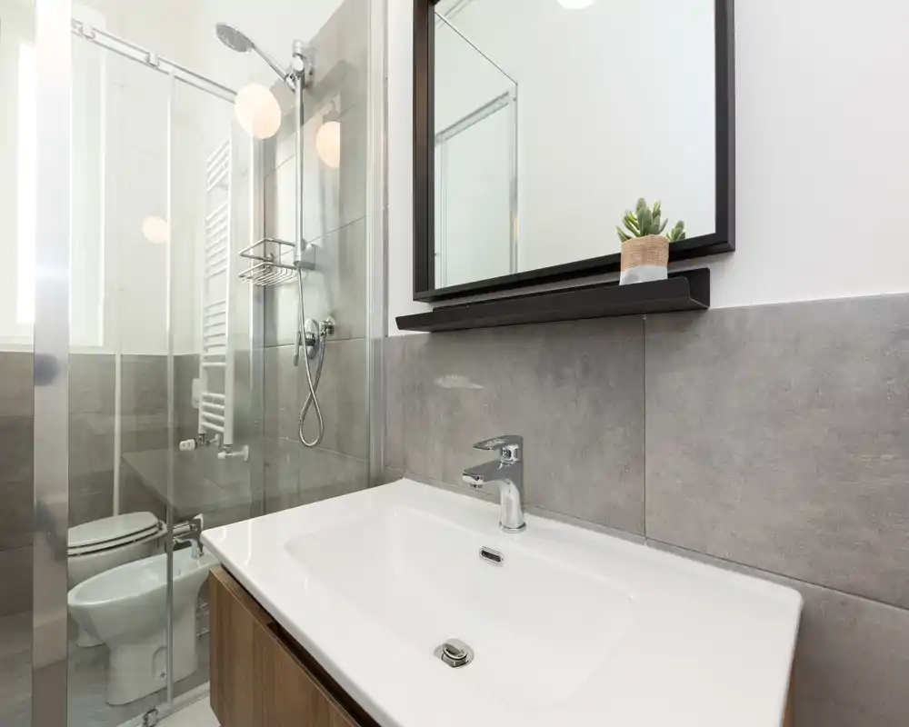 Bagno moderno e ben illuminato con lavabo bianco, rubinetto cromato, specchio a parete e doccia con parete in vetro e soffione: design pulito e contemporaneo, ideale per annunci.