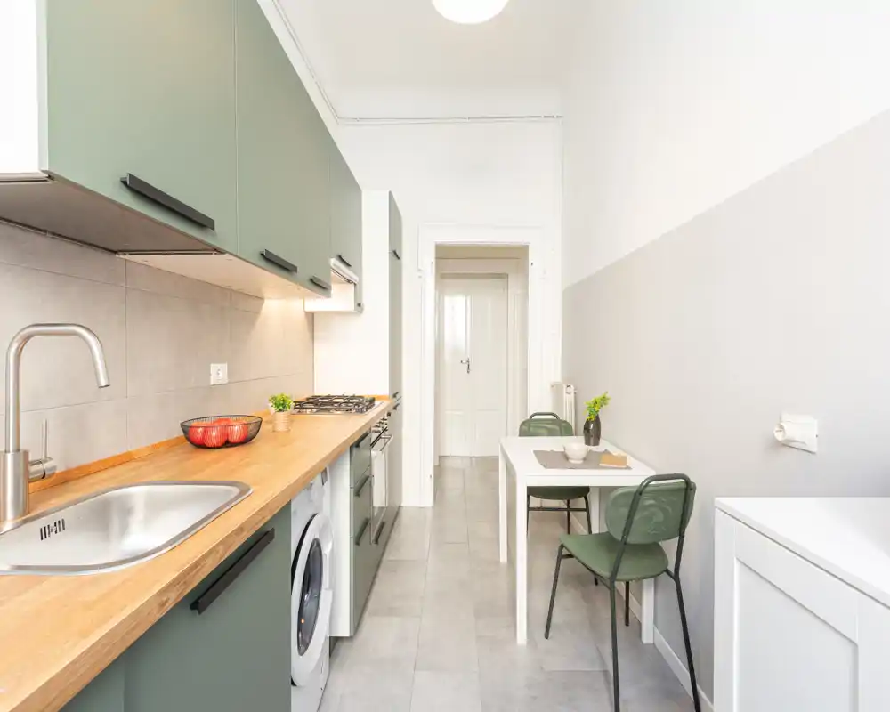 Cucina moderna a corridoio luminosa con mobili verde oliva, piano in legno, elettrodomestici integrati e piccolo tavolo da pranzo — funzionale ed elegante per la vita urbana.