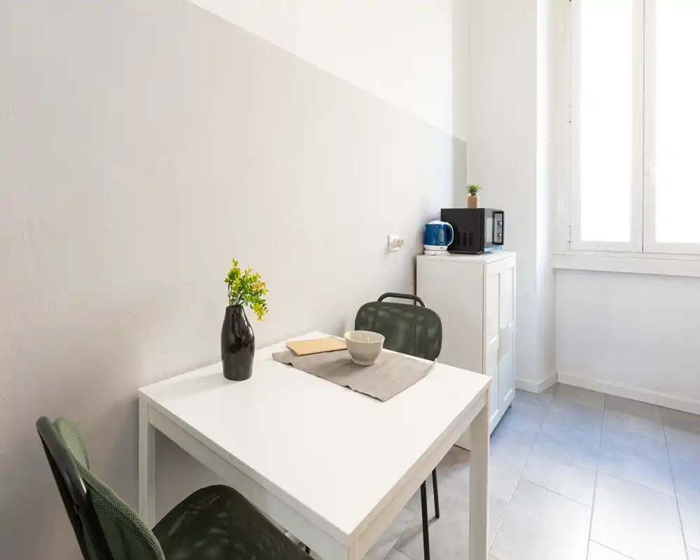 Angolo pranzo luminoso e minimal con tavolo bianco, due sedie e un piccolo mobiletto; la luce naturale dalla finestra rende l’ambiente arioso.