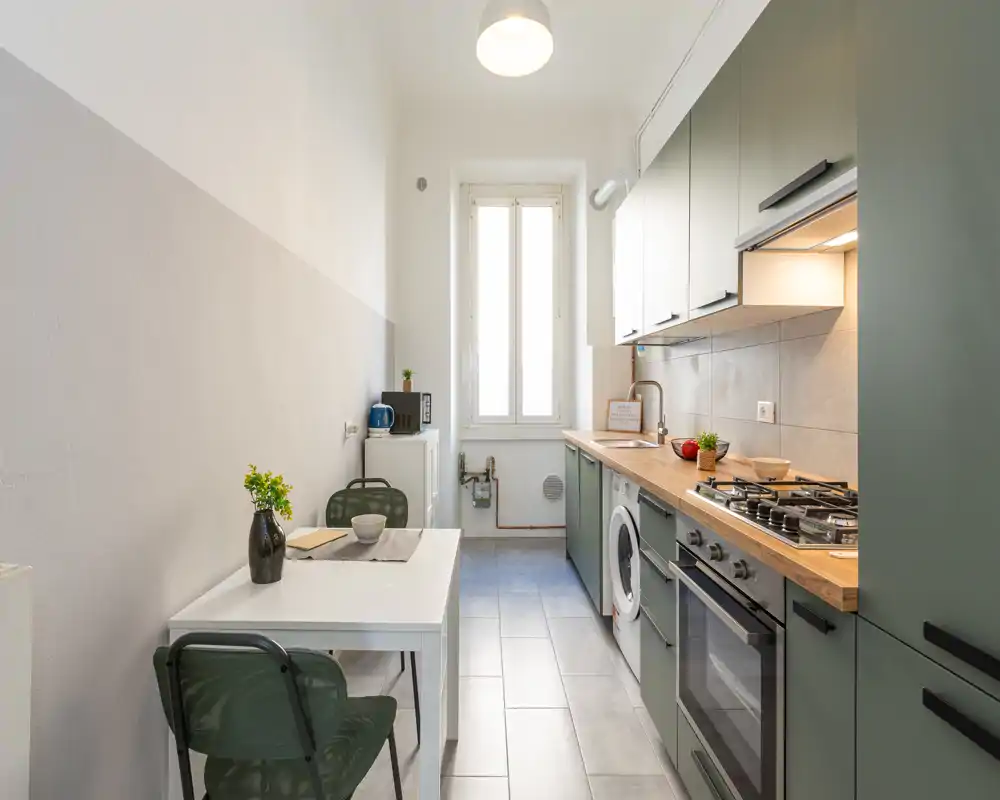 Cucina a corridoio luminosa con basi verdi, piano in legno, forno incassato e piccolo tavolo da pranzo — pratica, ben illuminata e arredata con gusto per spazi compatti.