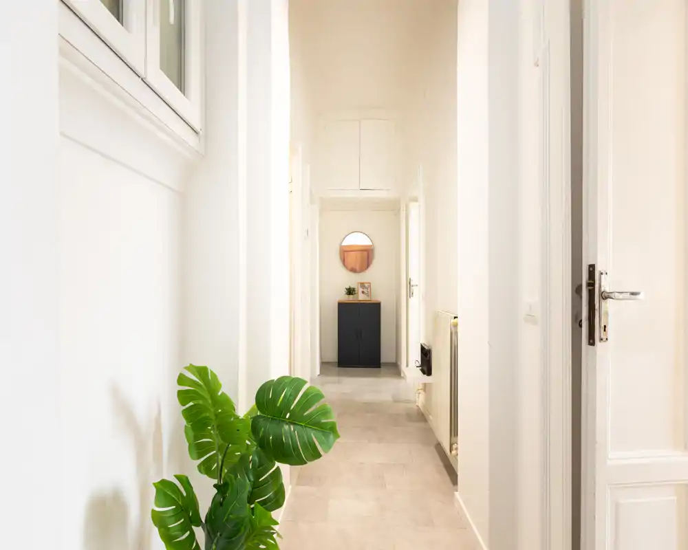 Ingresso luminoso e minimalista con pavimento chiaro in piastrelle, un piccolo mobile con specchio rotondo in fondo e una pianta decorativa in primo piano.
