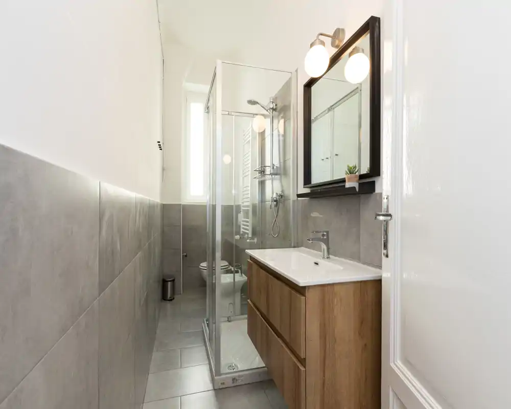 Bagno moderno e compatto con box doccia in vetro, mobile lavabo sospeso con cassetti in legno, specchio e luce naturale proveniente da una finestra stretta e alta.