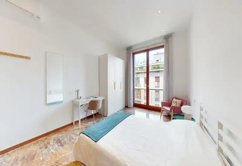 Bright, modern bedroom with a double bed, wardrobe, desk and balcony door letting in abundant natural light — clean lines and soft accents create a calm, comfortable space.