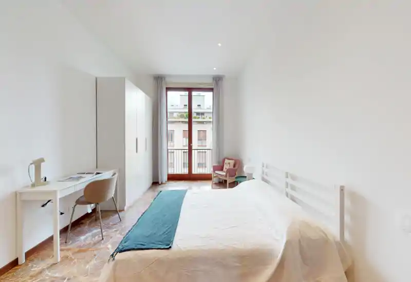 Camera da letto luminosa e moderna con letto matrimoniale, scrivania e armadio di fronte alla porta del balcone. Stile minimalista pulito, luce naturale e una comoda poltroncina vicino alla finestra.