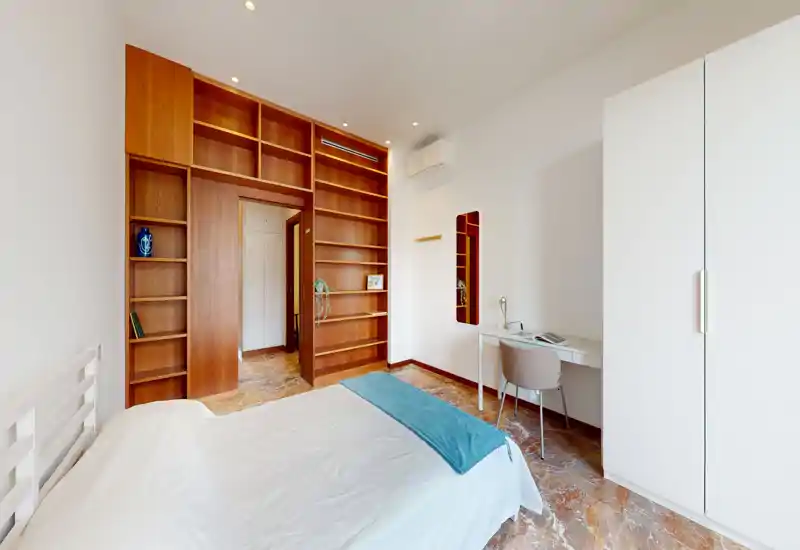Bright, modern bedroom with a comfortable double bed, white linens and a blue throw. Built-in wooden shelving and a small desk create a functional, stylish sleeping and working area.