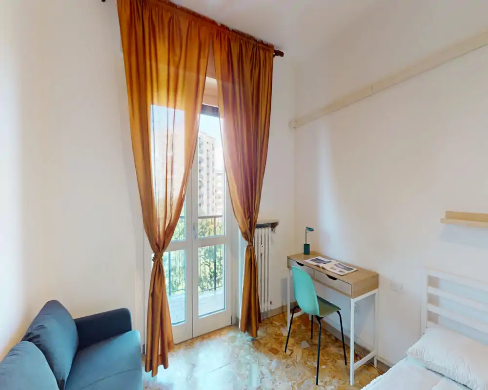 Dormitorio luminoso con gran ventana y cortinas cálidas; cama individual, pequeño escritorio y sofá crean un espacio funcional y bien iluminado, ideal para estudiante o persona sola.