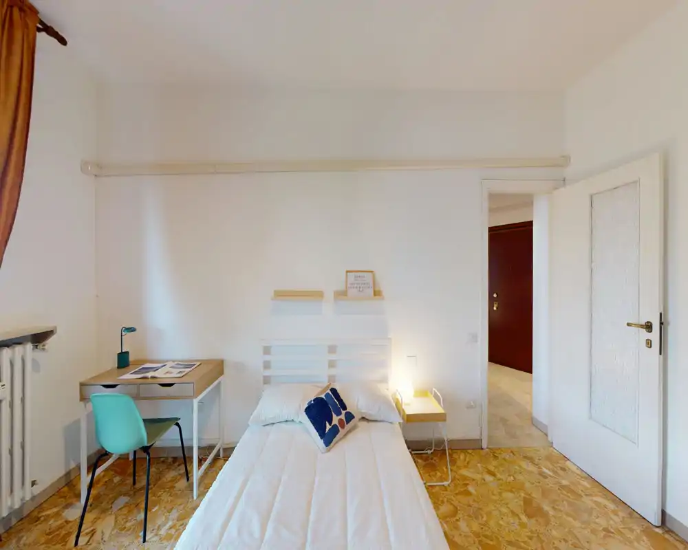 Dormitorio luminoso y minimalista con cama doble centrada contra una pared blanca, una mesita con lámpara y un escritorio compacto junto a la ventana — limpio, luminoso y funcional.