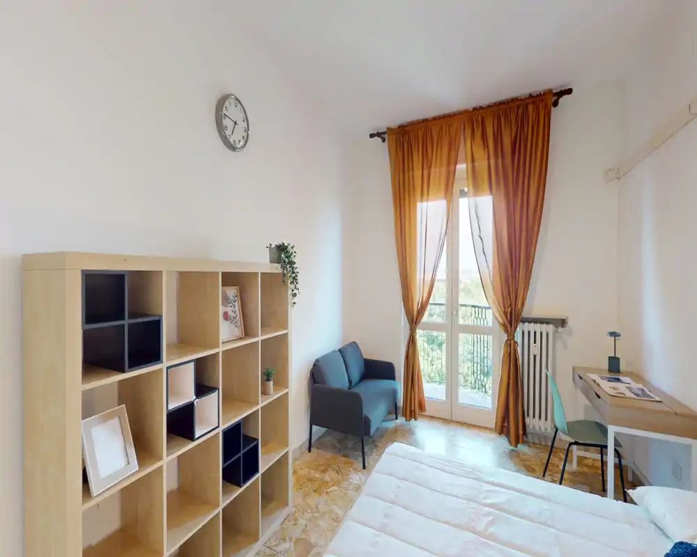 Dormitorio luminoso y bien presentado con cama visible, un pequeño sofá junto a la ventana, estantería tipo cubo y escritorio — cortinas cálidas y luz natural generan un ambiente acogedor.