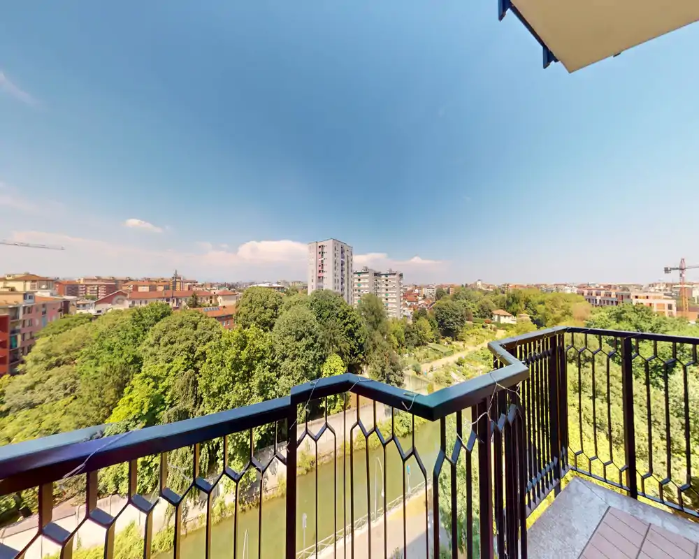Balcón soleado con barandilla metálica que domina un canal con árboles y edificios de la ciudad; amplia vista panorámica y cielo despejado, perfecto para un área exterior.