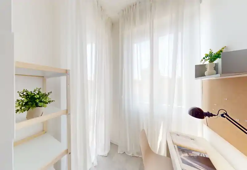Bright, compact home office corner with a white desk, reading lamp, cork noticeboard and sheer curtains that let in soft natural light.