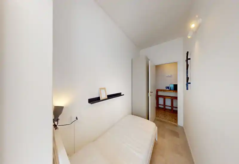 Compact single bedroom with a neatly made bed, wall-mounted shelf and soft lighting. The room is clean, simply furnished and well-lit, ideal for a short stay or a budget rental.