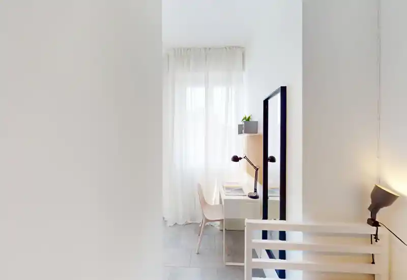 Compact bedroom corner with a small desk by a sheer-curtained window, bright natural light and simple Scandinavian-style furnishings including a bedside/bed slat visible in the foreground.