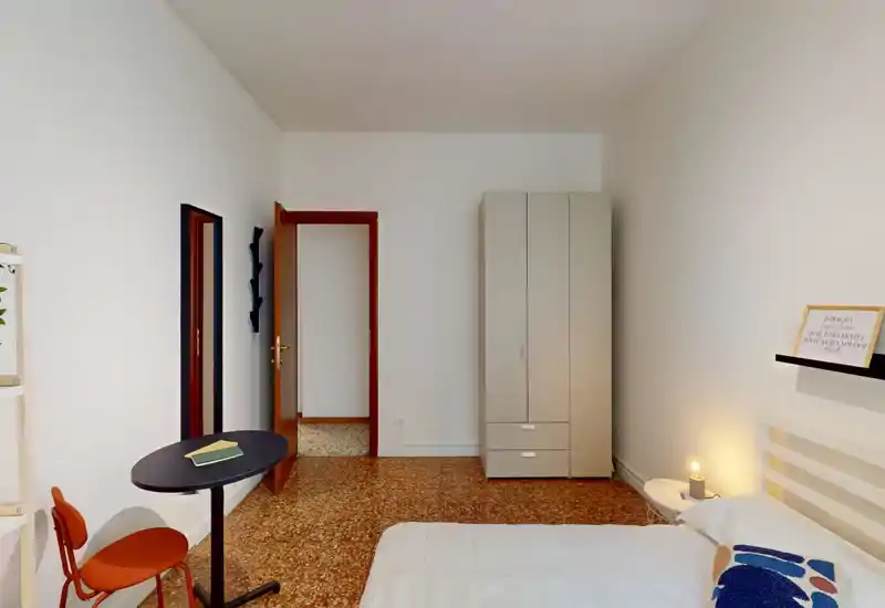 Bright, simply furnished bedroom with a double bed, bedside table and wardrobe; cozy lamp and small workspace create a comfortable, well-lit space ideal for short-term stays.