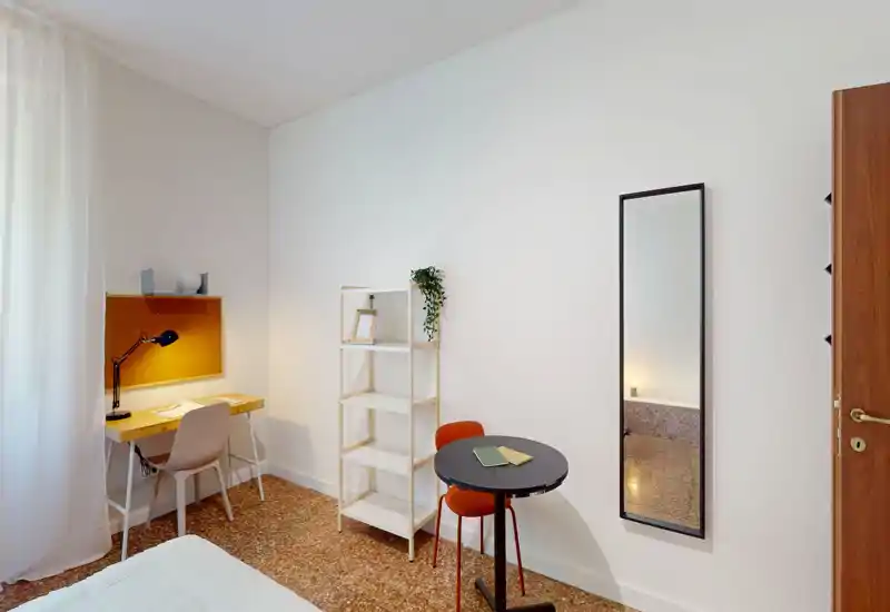 Bright, minimalist bedroom corner with a visible bed, a small desk with lamp, shelving and a round table — ideal for single stays or work-and-rest setups.